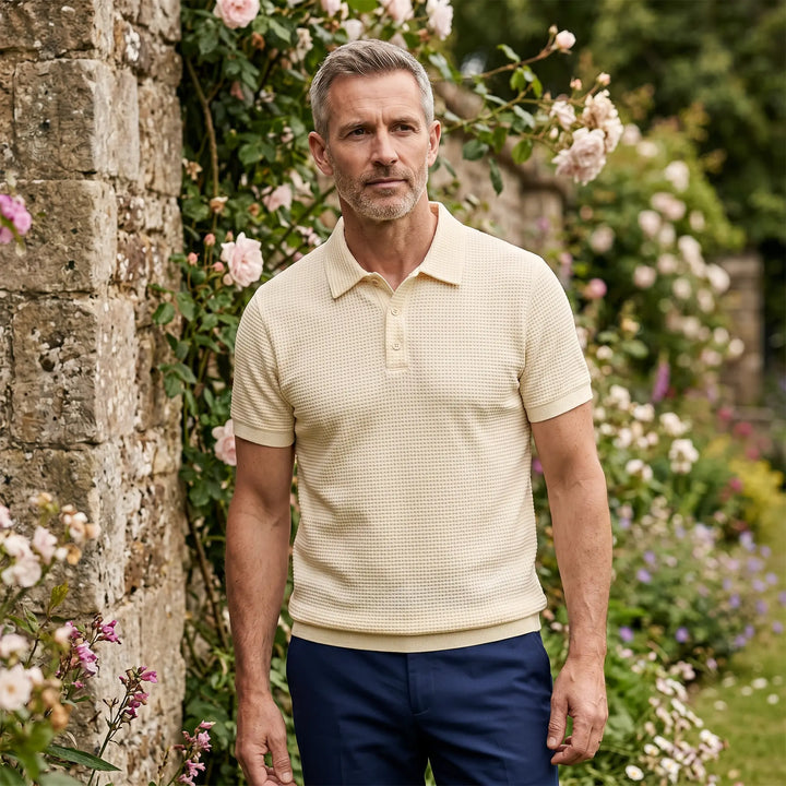 A distinguished man in a light yellow textured knit polo shirt and navy trousers stands in a garden setting, with a stone wall and blooming roses providing a refined backdrop.
