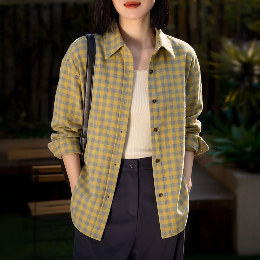 A woman models a relaxed-fit yellow and grey plaid brushed cotton shirt with a POLO collar, paired with a cream tank top and dark trousers. She wears a black shoulder bag, showcasing a casual yet chic outdoor lifestyle look. The shirt features button-down front and cuffed sleeves.