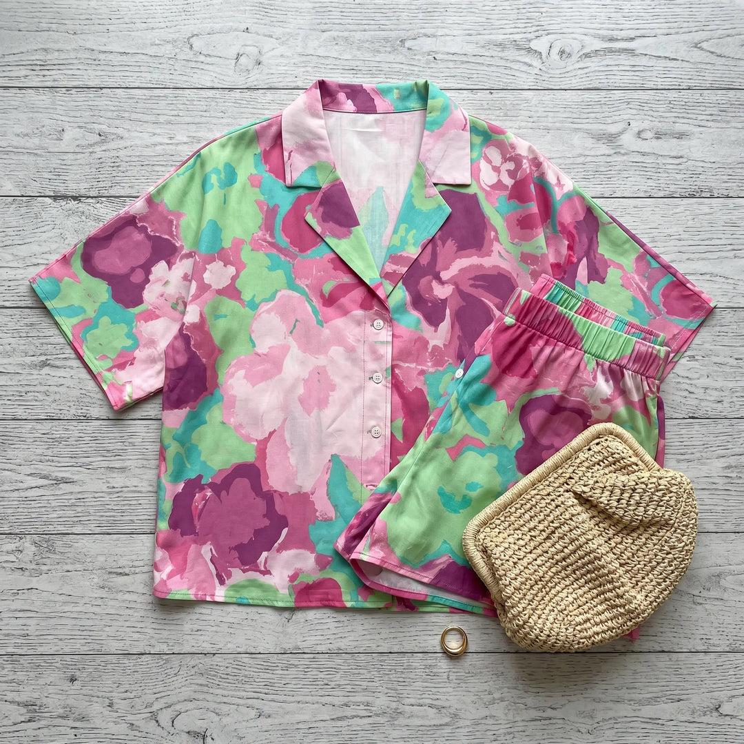 Aevoria abstract floral print loungewear set featuring a camp-collar shirt and elasticated shorts in vibrant fuchsia, magenta, and mint green against a light wooden background.