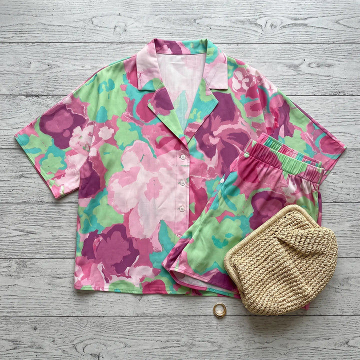Aevoria abstract floral print loungewear set featuring a camp-collar shirt and elasticated shorts in vibrant fuchsia, magenta, and mint green against a light wooden background.