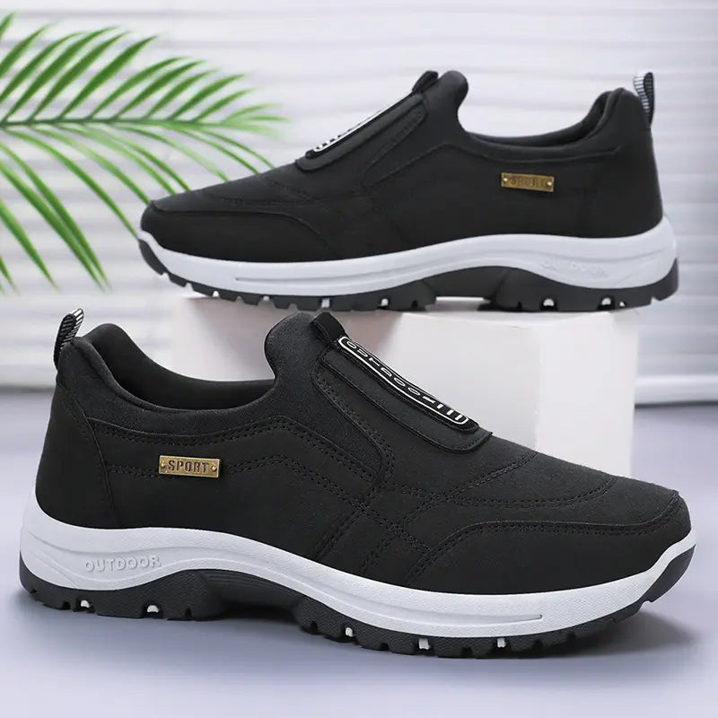 Aklos men's black slip-on casual walking shoes shown from the side, featuring a breathable textile upper, white EVA midsole, and black lugged rubber outsole with 'OUTDOOR' text detail.