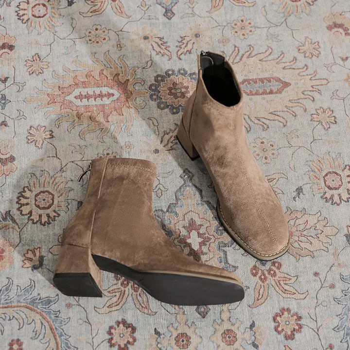 A pair of chic, light brown suede-like ankle boots with square toes and block heels, shown from a top-down angle on a patterned rug.