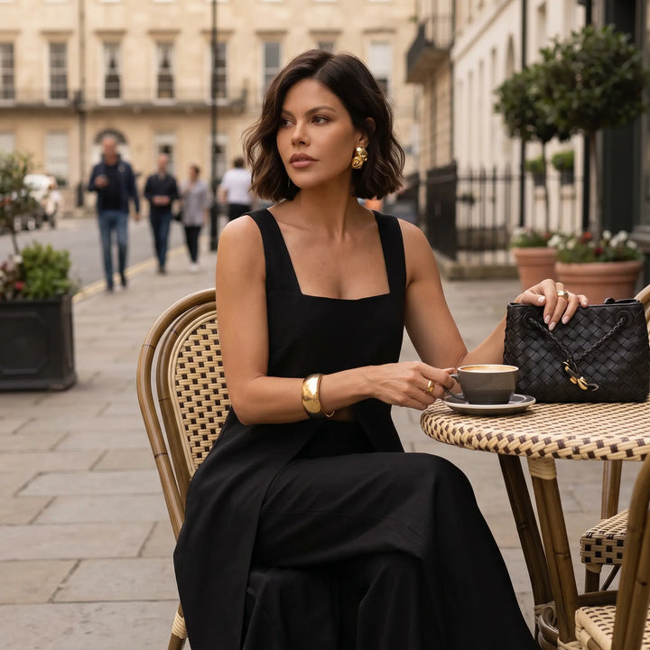 Model wearing a black Arvyla sleeveless maxi top and wide-leg trousers, sitting at a cafe table with a coffee cup, showcasing elegant, sophisticated style.
