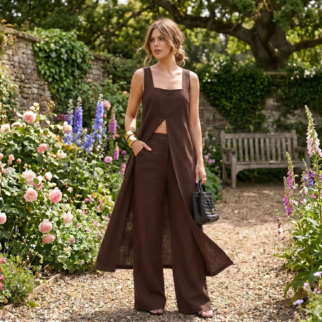 A woman models the Arvyla chocolate brown linen blend two-piece set featuring a sleeveless maxi top with a front split and wide-leg trousers, standing in a garden setting with flowers.
