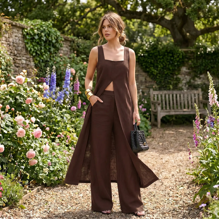 A woman models the Arvyla chocolate brown linen blend two-piece set featuring a sleeveless maxi top with a front split and wide-leg trousers, standing in a garden setting with flowers.