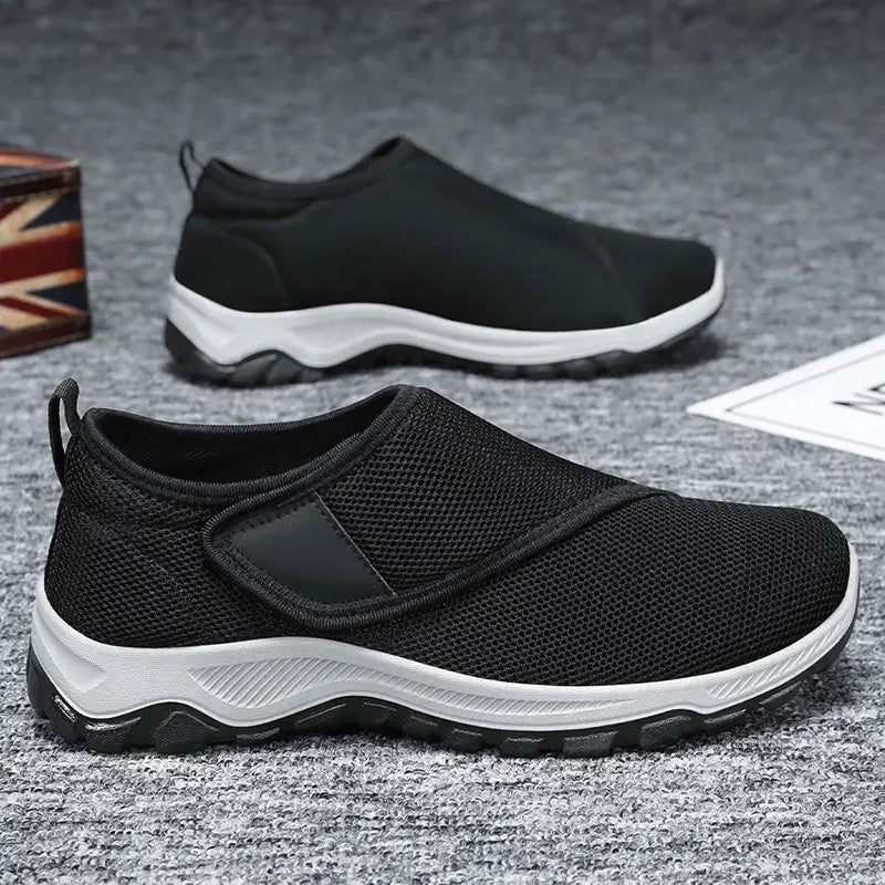 Pair of black men's breathable mesh slip-on sneakers with adjustable hook-and-loop strap, white EVA midsole, and black lugged rubber outsole displayed from the side.