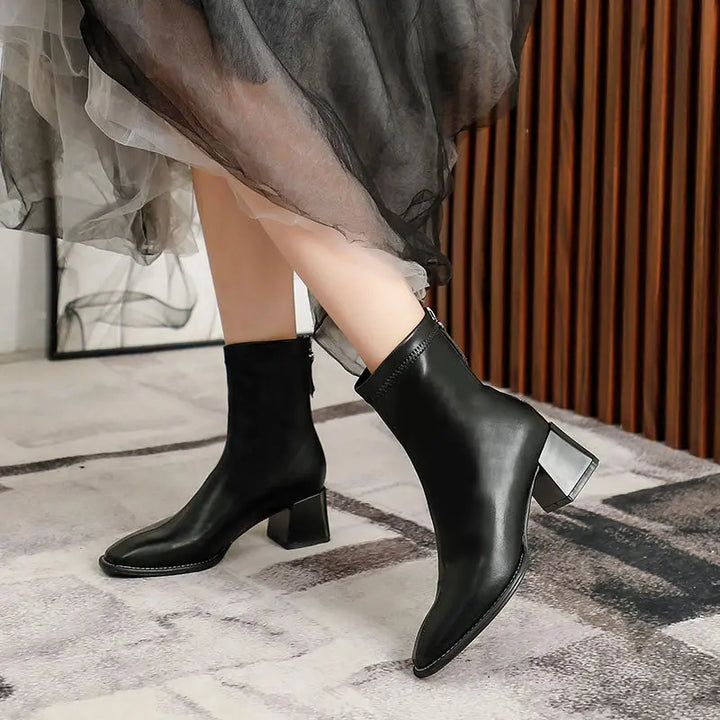Black faux leather ankle boots with a square toe, block heel, and rear zipper, worn by a model with a flowing skirt.