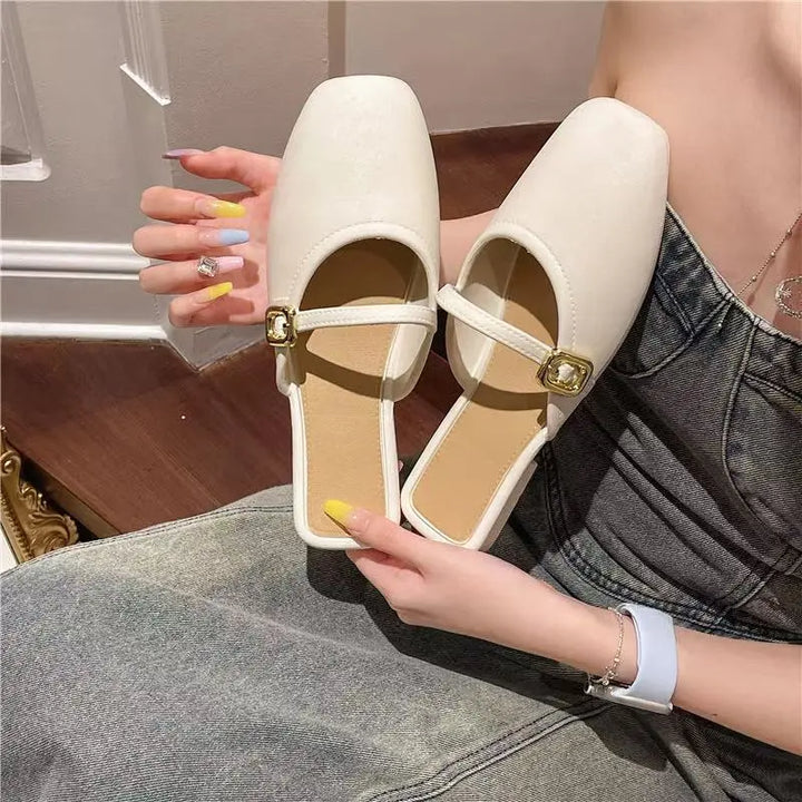A pair of ivory Azoria square-toe mules with gold-tone buckle straps are showcased by hands against a backdrop of denim clothing.
