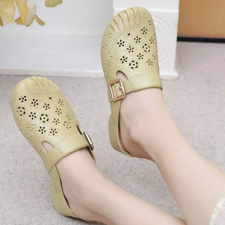 Close-up of light beige textured synthetic mules with an embossed geometric pattern, worn on feet, featuring a gold-tone adjustable buckle strap and a round toe.
