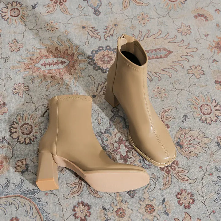 Beige faux leather ankle boots with a square toe, chunky block heel, and back zipper, elegantly displayed on a patterned rug.