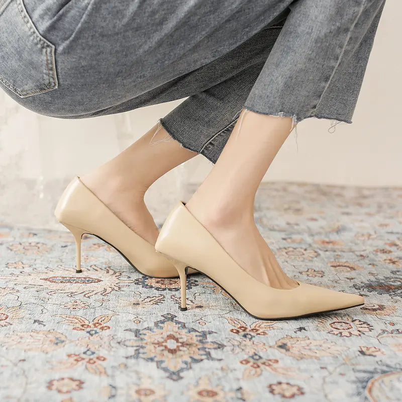 Close-up view of beige high-gloss faux leather stiletto heels with a pointed toe, worn by a model on a patterned rug, showcasing the elegant low-cut vamp and slender heel.