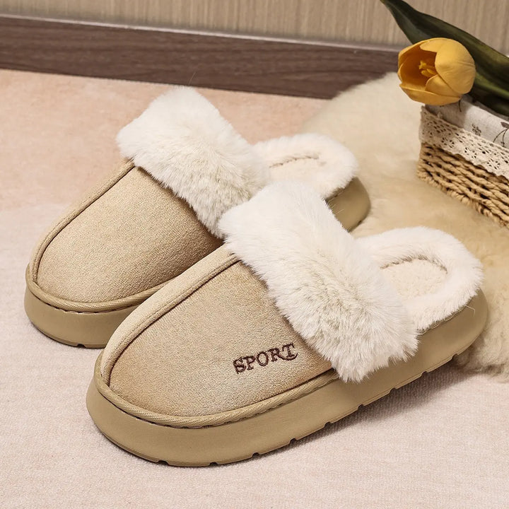 A pair of beige faux suede mule slippers with plush white faux fur trim and an embroidered "SPORT" logo on the vamp, displayed on a soft rug with decorative elements in the background.