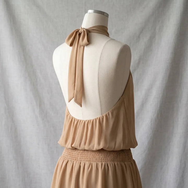 Back view of a flowing beige halter neck mini dress with a shirred waistband and an open back secured by a delicate tie closure.