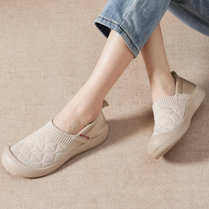 A woman wearing light beige knit slip-on sneakers with a diamond quilted pattern, paired with casual blue jeans, shown in a lifestyle setting on a textured floor.