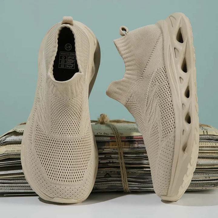 A pair of light beige, slip-on knit trainers with a textured upper and sculpted wave-patterned sole, shown from a three-quarter angle on a stack of newspapers.