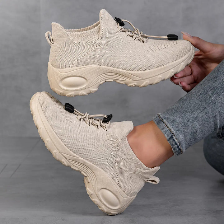 Beige engineered knit low-top sneakers with a quick-lace system, chunky white sole, and a subtle woven pattern, shown on feet and held by hands against a grey studio background.