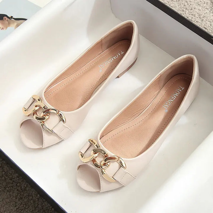 A pair of light beige peep-toe flat shoes with a gold-tone chain link embellishment across the vamp and a flat heel.