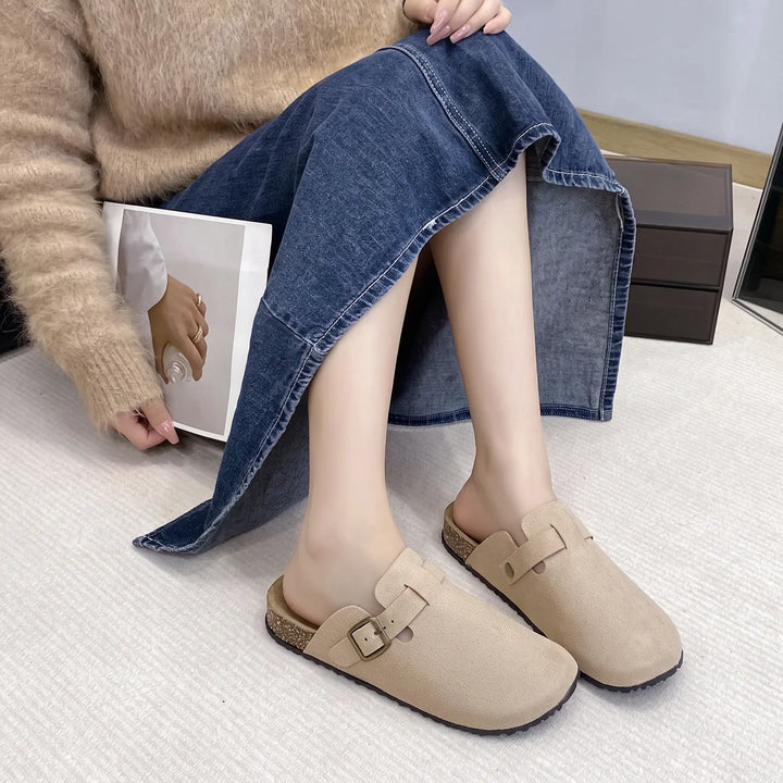 Lifestyle image of beige suede-like mules with an adjustable brass buckle, worn by a woman with a denim skirt and fuzzy sweater.