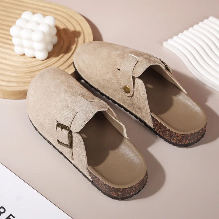 A pair of beige suede-like mules with an adjustable brass-toned buckle, showcasing the rounded toe, cork-like midsole, and plush faux shearling lining.
