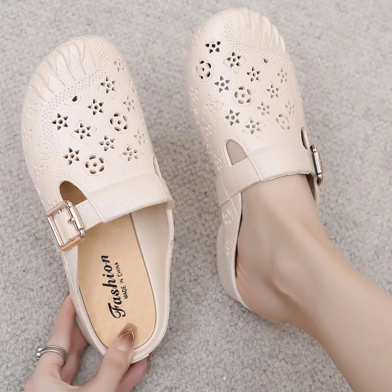 Close-up of beige textured synthetic mules with embossed pattern and adjustable gold buckle strap, shown on a foot with the &