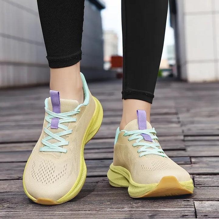 Pair of beige wide-fit athletic trainers with mint green and purple accents, worn on feet with black leggings, displayed on a wooden surface.