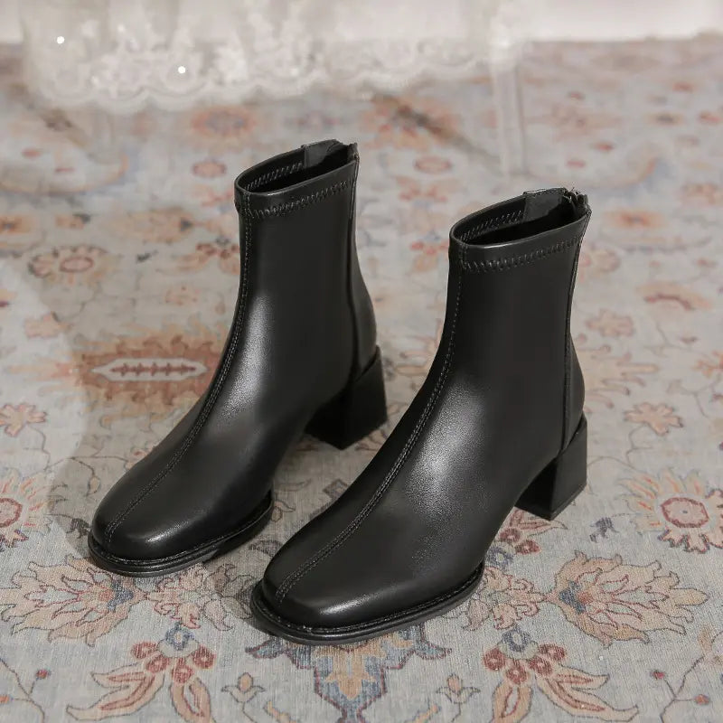 Black faux leather ankle boots with a square toe, chunky block heel, and back zipper closure, presented on a patterned rug.