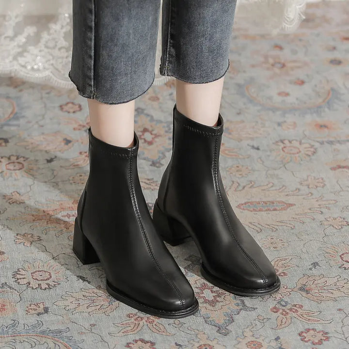 A pair of black faux leather ankle boots with a square toe and chunky block heel, worn with cropped jeans on a patterned rug.