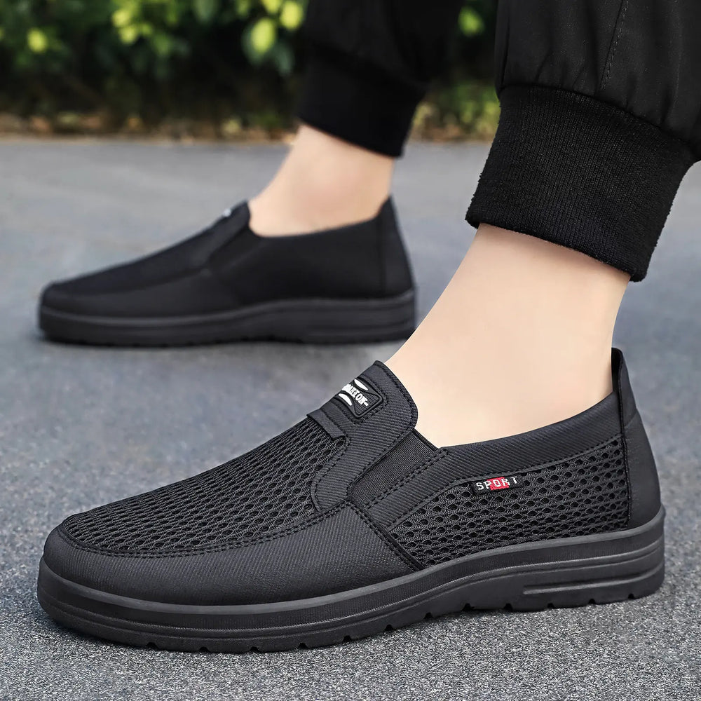 Men's black breathable mesh slip-on loafers worn outdoors, featuring elasticated sides and a black PVC sole with a subtle 'SPORT' logo detail on the side.