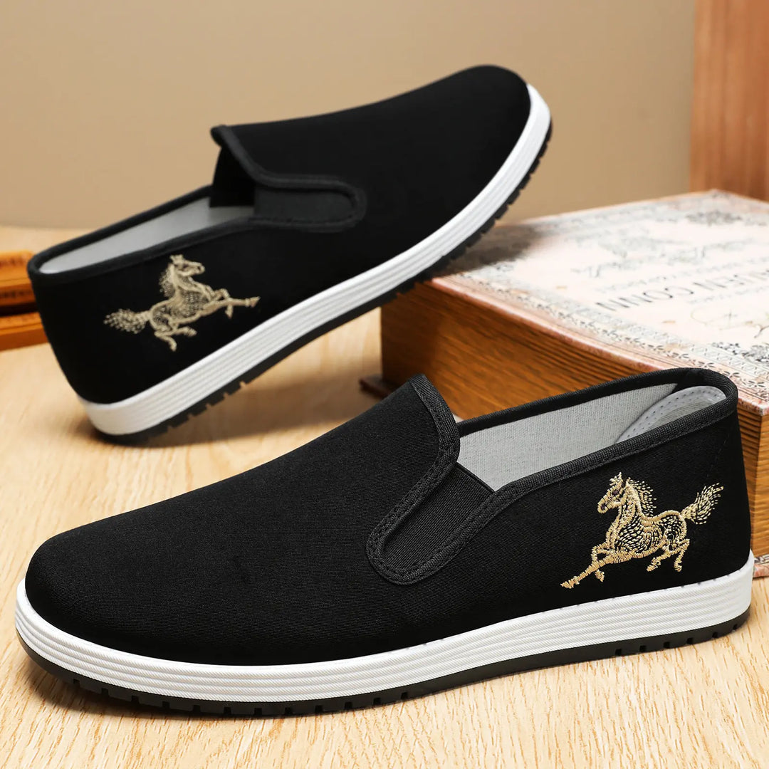 Men's black canvas slip-on loafers featuring intricate gold horse embroidery on the side and a contrasting white rubber sole, shown from a three-quarter front angle.