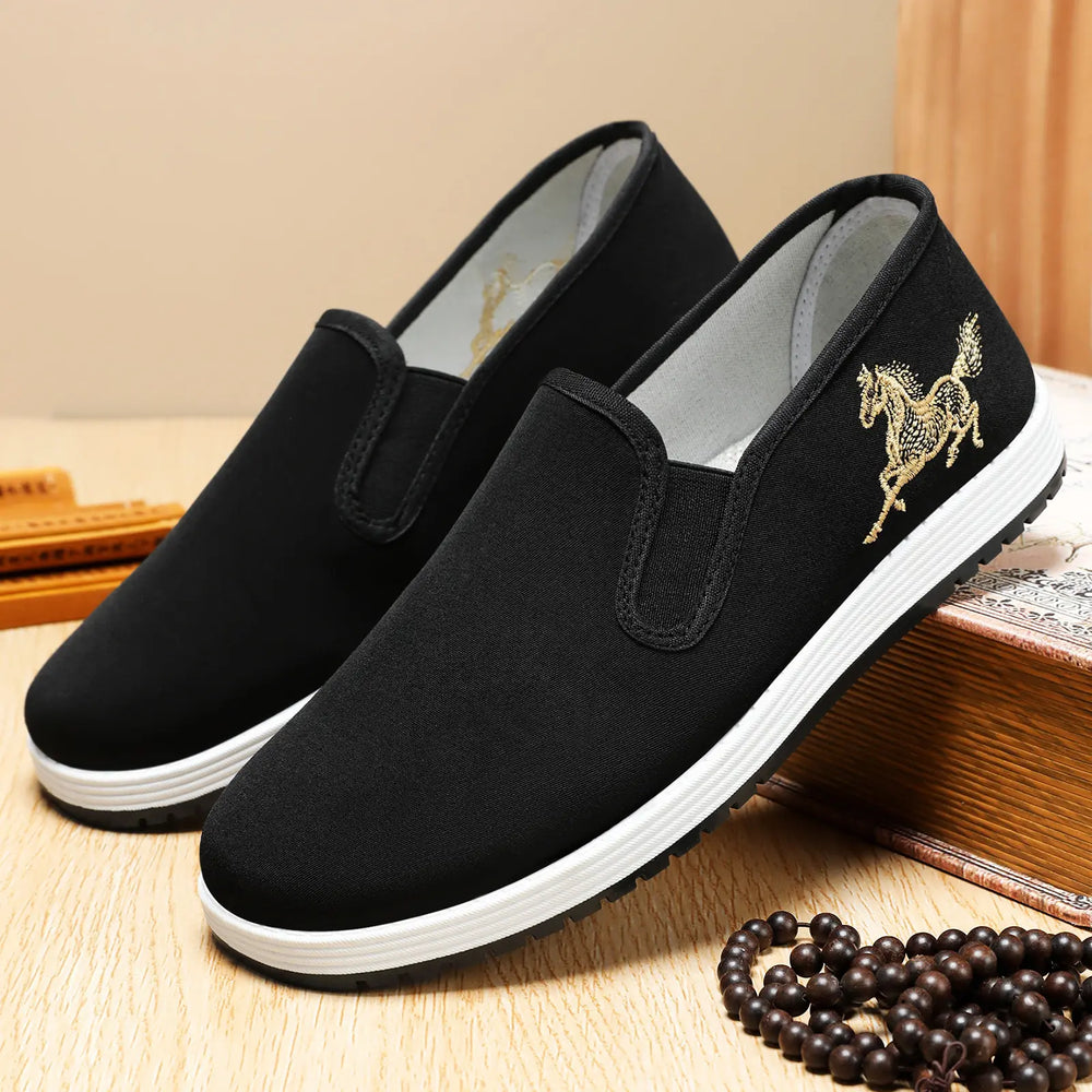 Men's black canvas slip-on loafers featuring intricate gold horse embroidery on the side and a white rubber sole.