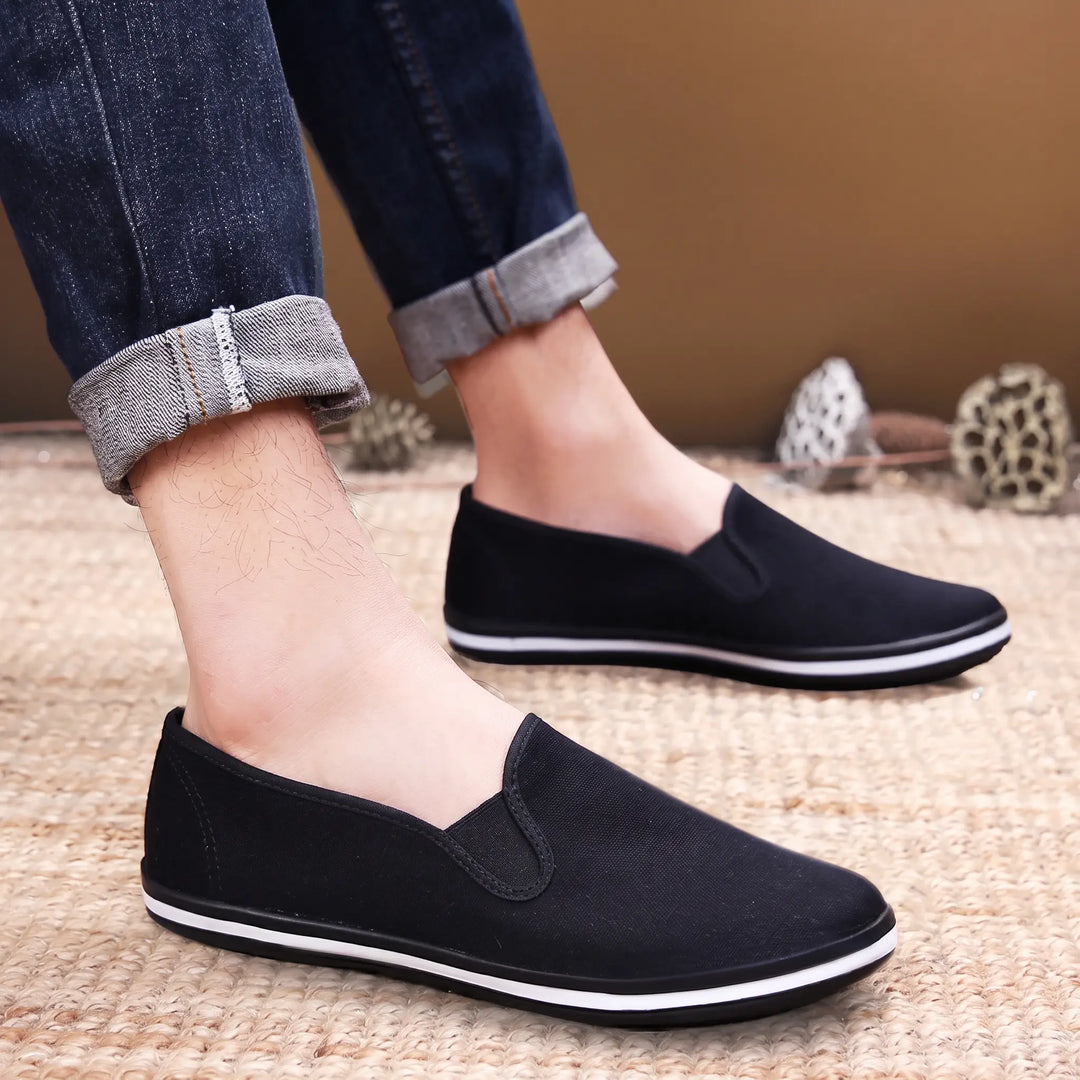 Black canvas slip-on loafers worn by a person, featuring a round toe and a white stripe around the sole edge, set against a textured woven background.