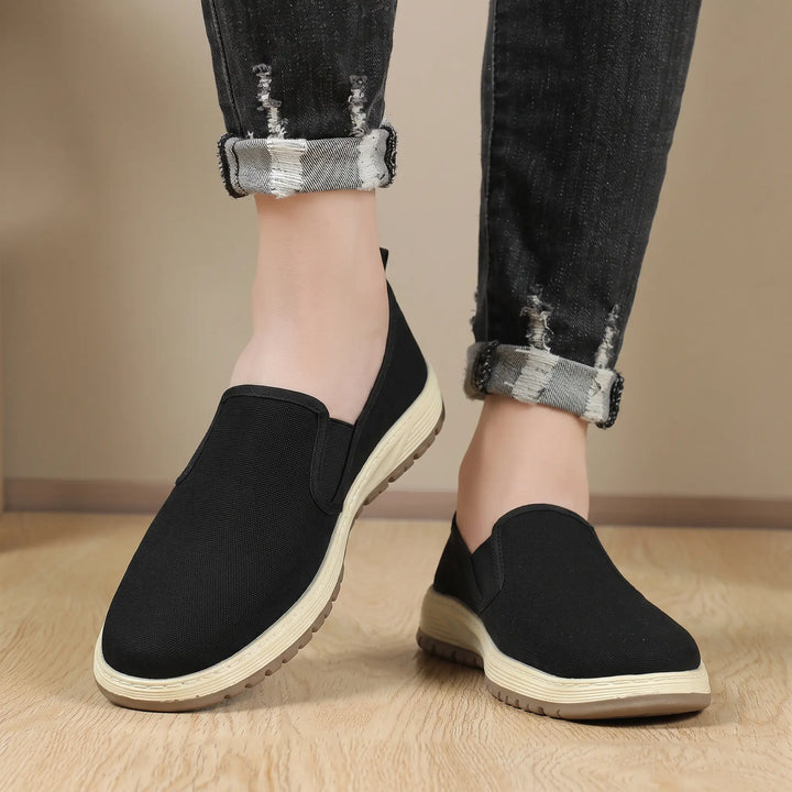 A person wearing black Zentia slip-on canvas shoes with cuffed distressed jeans, showcased from a three-quarter angle against a textured neutral background.