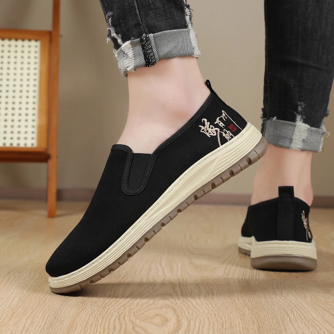 Black canvas slip-on shoes with subtle embroidered detail on the heel, shown on feet with distressed jeans, offering a comfortable and stylish casual look.