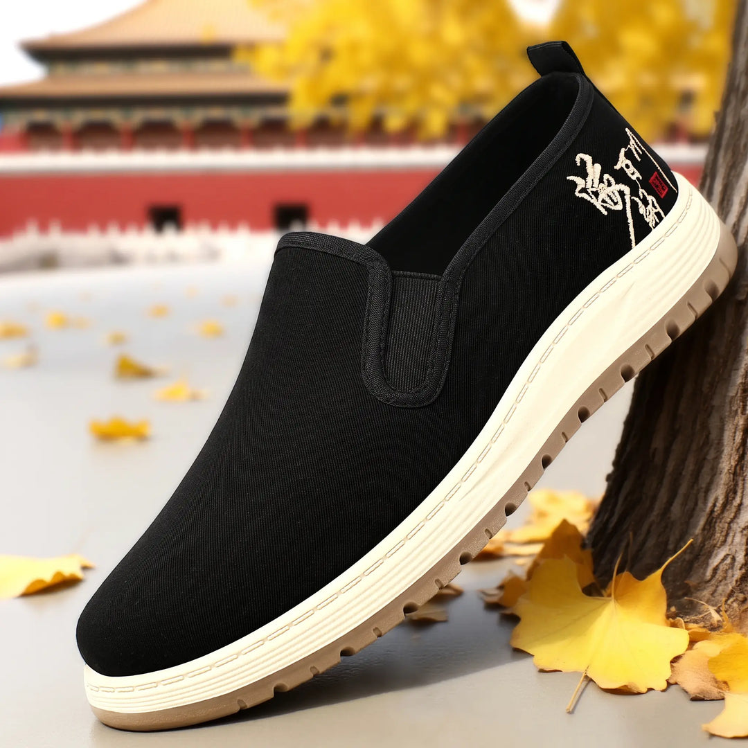 Black canvas slip-on shoes with a round toe, elastic side gores, and subtle embroidered detailing on the heel, shown from a three-quarter angle.