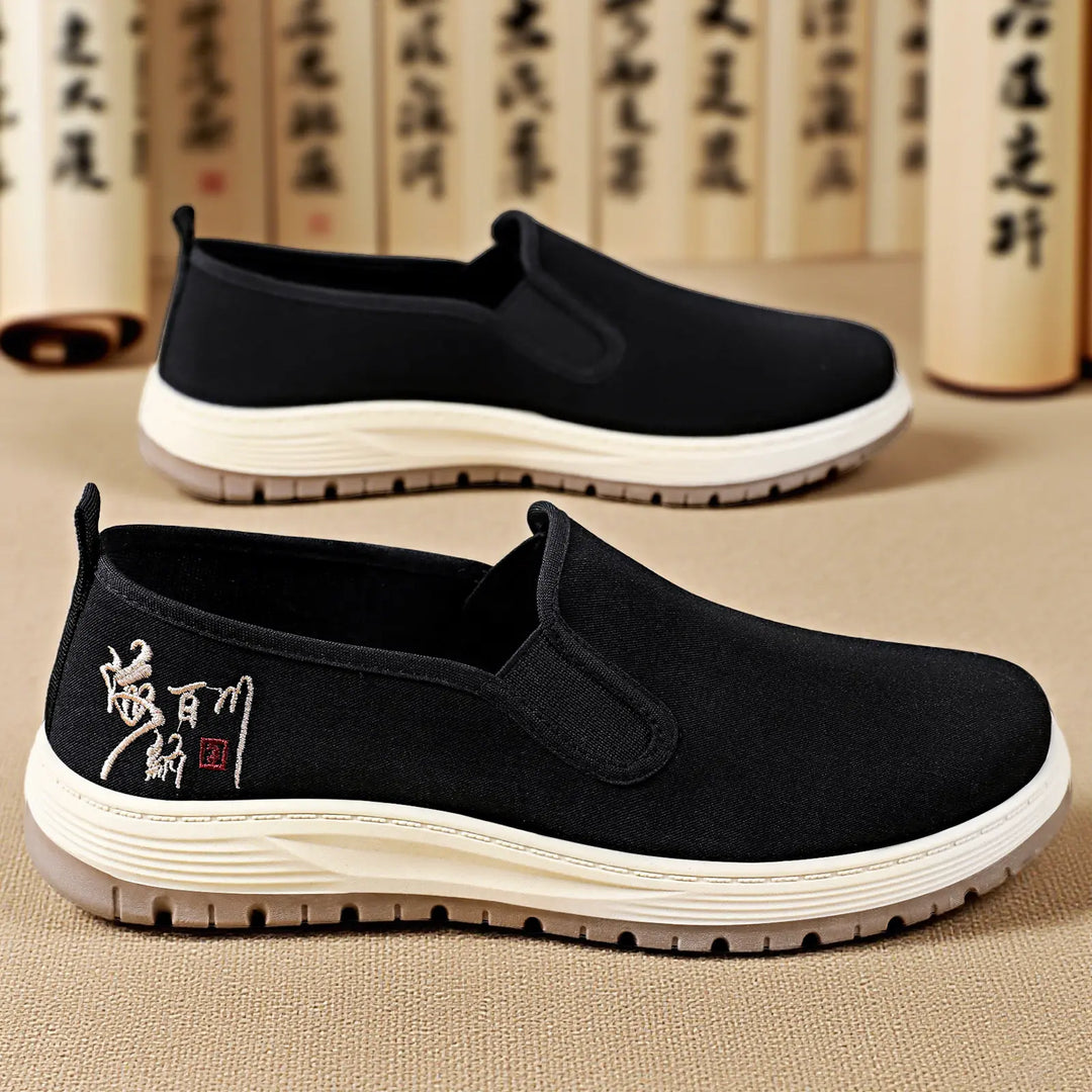 A pair of black canvas slip-on shoes with a subtle embroidered emblem on the lateral heel, viewed from a three-quarter angle against a textured background.