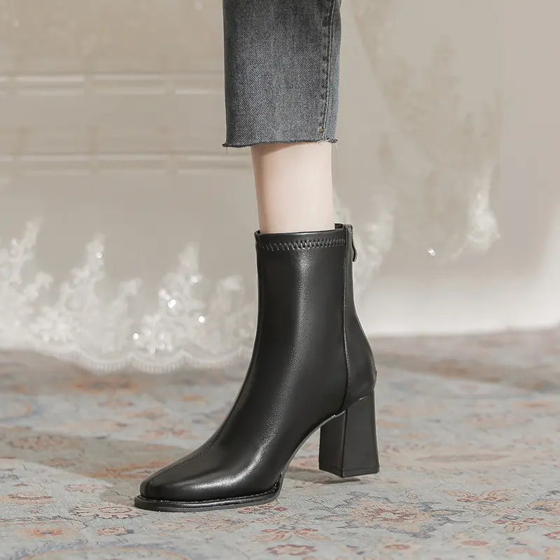 Black faux leather ankle boots with a chunky block heel and square toe, worn with grey cropped jeans, showcasing the back zipper detail.
