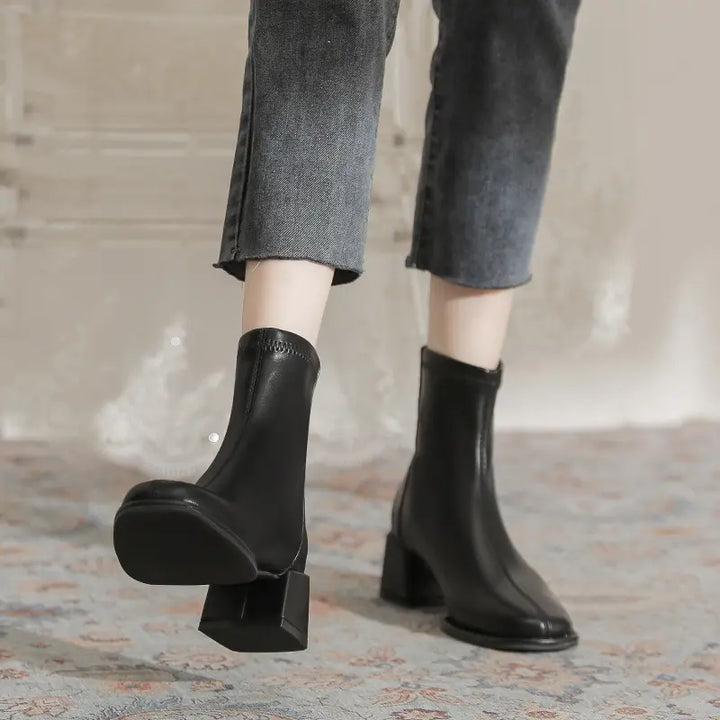 A pair of black faux leather ankle boots with chunky block heels and a square toe, worn by a person in cropped grey jeans.