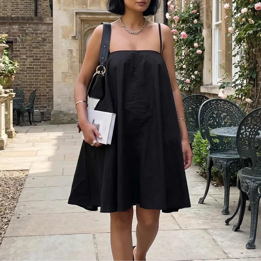 A woman walks outdoors wearing a black cotton A-line mini sling dress with thin adjustable spaghetti straps and a square neckline, styled with a necklace and handbag.