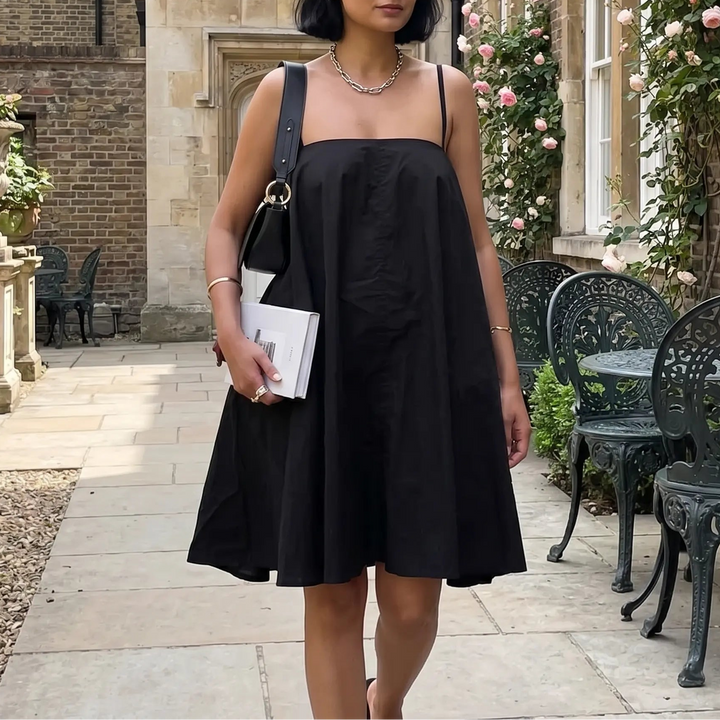 A woman walks outdoors wearing a black cotton A-line mini sling dress with thin adjustable spaghetti straps and a square neckline, styled with a necklace and handbag.