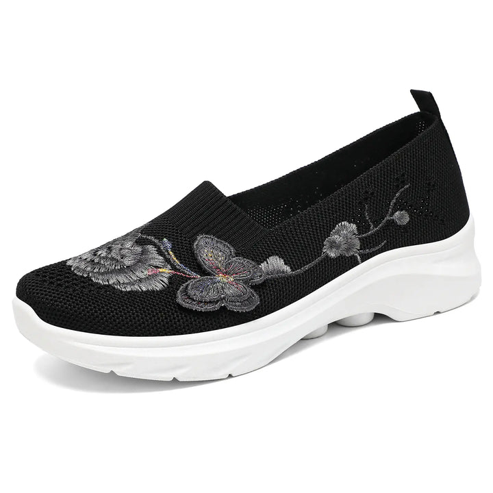 Black embroidered knit slip-on sneakers featuring delicate floral and butterfly motifs, a comfortable round toe, and a white cushioned sole.