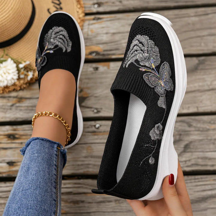 A pair of black embroidered knit slip-on sneakers with intricate floral and butterfly motifs, shown on a woman&