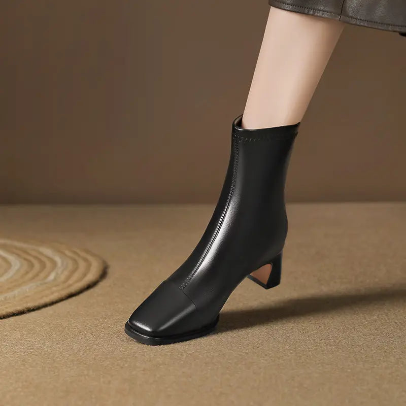 Black faux leather ankle boots with a square toe and block heel, worn by a model showing the mid-calf shaft in a subtle lifestyle setting.