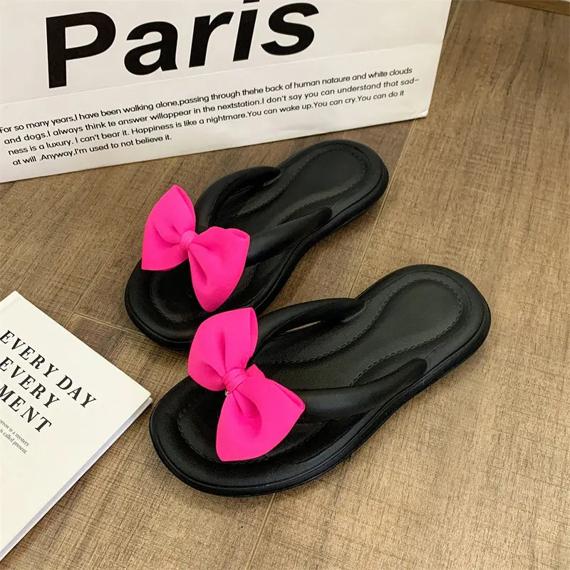 Black flip-flops with vibrant fuchsia pink bows and gold teddy bear charms, displayed on a wooden surface next to books.