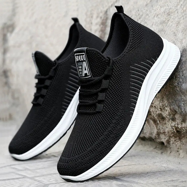Close-up three-quarter view of black breathable knit mesh men's sneakers with white EVA midsole and textured black rubber outsole. Features flat black laces and a 'SPORT 2028' label on the tongue.
