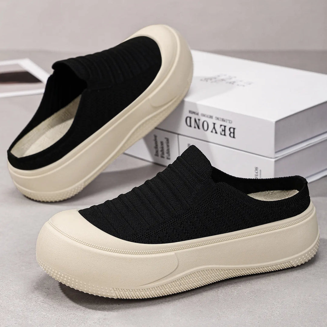 Black knit slip-on platform mules with a thick, creamy beige sole, shown from a three-quarter angle on a studio surface with books in the background.