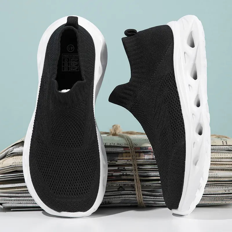 Pair of black knit slip-on sneakers with white sculpted wave soles, shown from front and three-quarter angles on a studio background.