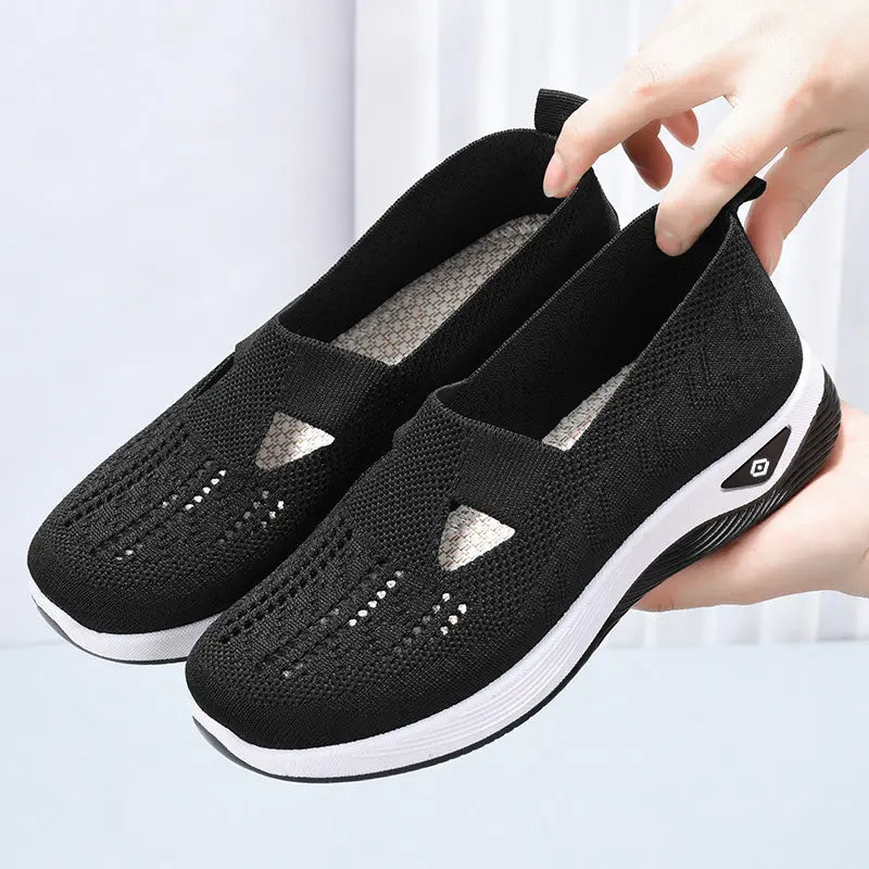 A pair of black breathable knit slip-on sneakers with white soles are shown from a three-quarter angle, held by hands against a soft studio background.