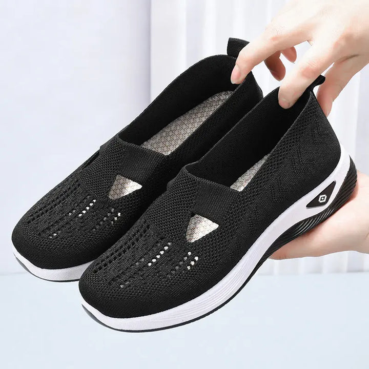 A pair of black breathable knit slip-on sneakers with white soles are shown from a three-quarter angle, held by hands against a soft studio background.