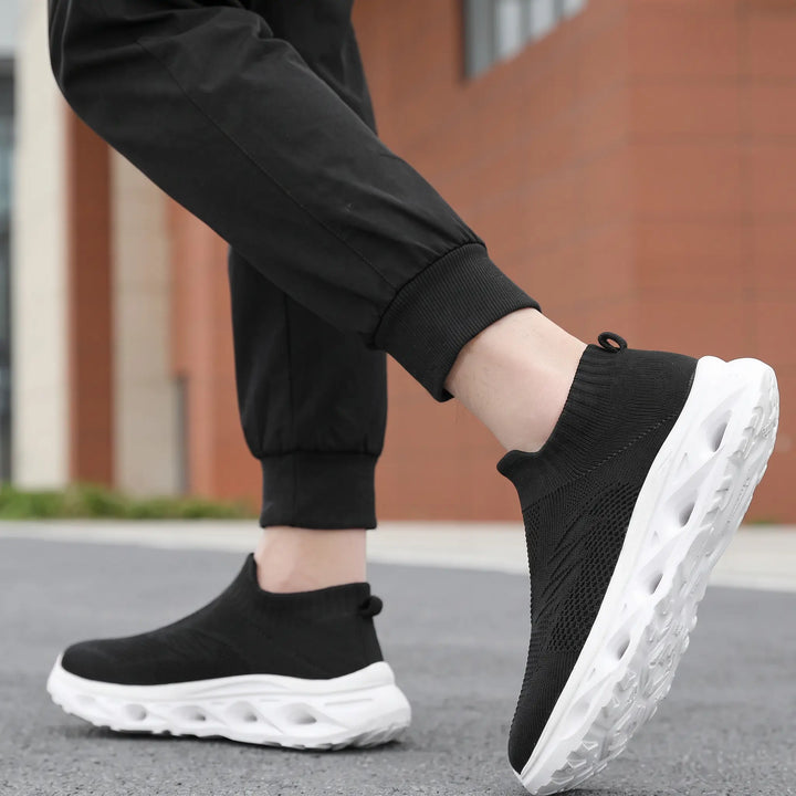Black knit slip-on trainers with white sculpted soles worn by a person in a casual outdoor setting.