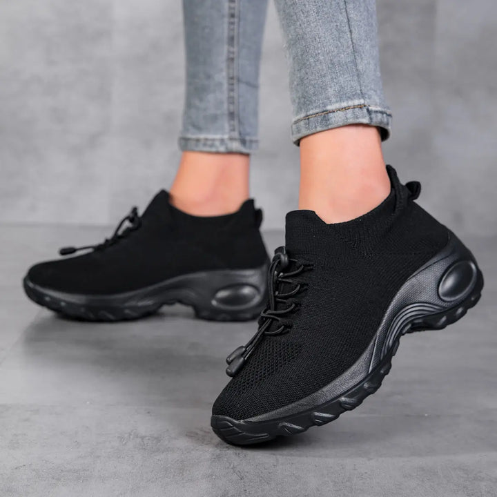 Black knit low-top sneakers with a sock-like fit, quick-lace system, and chunky white sole, shown worn on feet against a grey studio background.